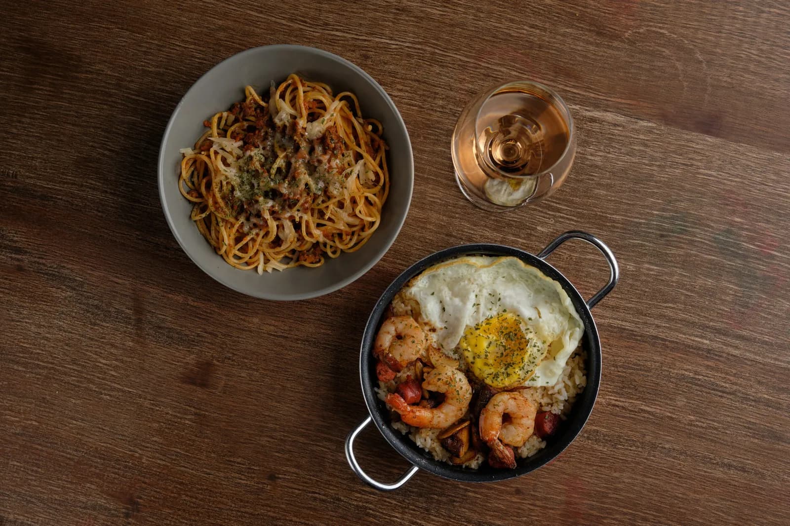 A pasta bowl, shrimp rice with a fried egg and a glass of rosé on a wooden table.