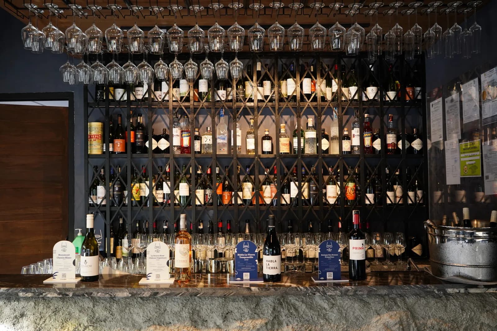 Project Vino bar backed by wine and spirits racks with rows of hanging glasses.