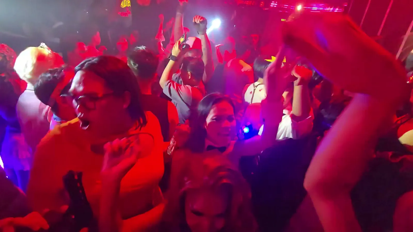 Packed dance floor under red club lights with guests raising their hands